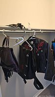 Black leather jacket and matching chaps hanging on hangers showing rose motif embroidery and silver decorative buttons with fringe. Two black leather purses hanging with fringe detail visible. Black leather gloves placed on closet shelf above.