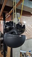 Three motorcycle helmets hanging by ropes in a basement, showing from angled side views: a Harley Davidson matte black helmet, a ZR helmet, and a CL-5 helmet.