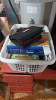 White laundry basket filled with puzzle boxes, travel Scrabble game in black case on top, basement setting