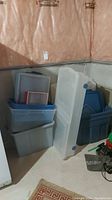 Gray and blue Rubbermaid storage totes stacked against wall with lids and clear under bed storage tote leaning on them. Located in basement with pink insulation on wall behind.