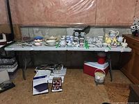 Full view of white plastic folding table with metal legs, placed in unfinished basement area with various household items on and around it.