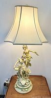 Full view of the 44.5 inch tall table lamp lit, showing the ceramic base with two sculpted women and the large cream-colored fabric shade.