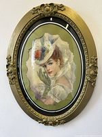 Clear front angle view of the oval gilt frame with the painting inside, showing the woman with a hat and gloves, backed by green matting.