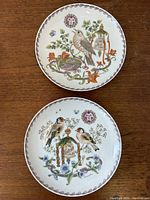 Pair of 9-inch diameter porcelain wall charger plates showing intricate bird and floral designs with detailed borders, photographed on a wooden surface.