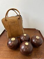 Four wooden lawn bowling balls arranged in front of a tan vinyl carrying case on a wooden surface.