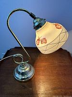 Photo of vintage desk lamp with curved metal arm and floral patterned glass shade on wooden surface, viewed from side.