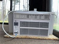 Front view of white Perfect Aire window air conditioning unit on a beige carpet near window. Unit has vent grills and attached cable.