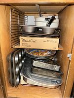 Baking pans, Pampered Chef mini loaf pan box, pizza stone, and round cake pans stacked inside a wooden cabinet.
