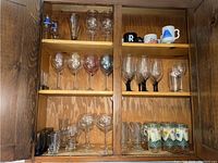 Cabinet with multiple shelves showing various stemware including wine glasses with floral designs, clear stemmed wine glasses with black base, water glasses with geometric pattern, and assorted mugs on the bottom shelf