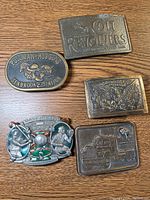 Overview of five different metal belt buckles showing various themes like Colt Revolvers, baseball, trucking, eagle emblem, and Pullman-Higgins Seabrook Station.