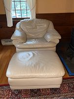 Front view of beige leatherette chair with matching ottoman, showing cushioned surfaces and stains.