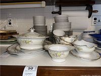 Wide shot of multiple stacks of white Empress china dinner plates, bowls, tea cups, saucers, serving bowls, and covered serving pieces with floral decoration and gold trim.