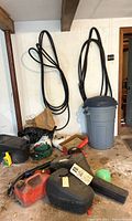 Wide view of garage corner showing hoses on wall, trash barrel, gas container, chainsaw cases and yard tools on floor.