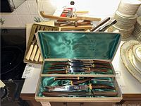 Vintage Sheffield steak knife set with wooden handles, displayed in green satin-lined case along with other kitchen items including plates and various cutlery