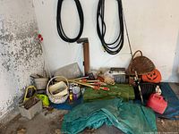 Cluster of garden and outdoor items including fishing rod, tarps, gas cans, hoses, baskets, and metal box against a wall.