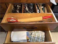 View of wooden drawer organizer containing assorted stainless steel flatware and kitchen gadgets like rolling pin and whisk; lower drawer contains kitchen towels and oven mitts.