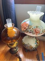 Photo showing two oil lamps side by side, one amber glass and brass, one cream floral painted glass shade with chimney.