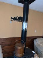 Metal wall art mounted on wall above wood stove, showing horse and carriage silhouette in black metal along with Basketville wooden firkin bucket on stove.