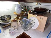 Wide view of corner kitchen counter showing Bodum hot chocolate pitcher, ceramic canisters, metal and glass mixing bowls, Corning Ware buffet server, glass serving platters stacked on box.