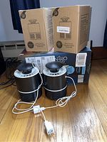 Two black and white photocatalytic mosquito killer lamps with power cords and plugs, alongside their original cardboard boxes on wooden floor