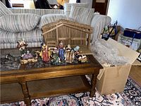 Full view of nativity scene figurines arrayed on a wooden table with stable in the background revealing a set of multiple characters and animals typical of a traditional nativity scene.