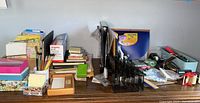 Photo showing a collection of office supplies including pens, pencils, organizers, stapler, glue, brushes, cards, tape, and notepads on a wooden desk.