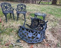 Wide shot showing two wrought iron chairs standing, and one loveseat upside down displaying intricate floral castings, placed on grass outdoors. Rust and damage visible on legs.