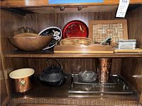 Shelf with assortment of serving items including wooden bowl, glass trays, copper bucket, teapot, coasters, trivets.