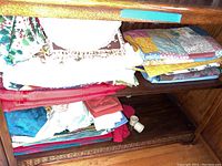 Shelf with assorted folded tablecloths, napkins, and placemats including patchwork quilted and solid colored fabrics in red, cream, and floral patterns.