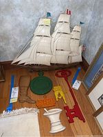 Photo showing a large off-white sailing ship model with three masts, multiple sails, and flags atop the masts. Surrounding the ship model on the floor are assorted colored wall plaques in different shapes and sizes including a brown boot, green round plaque with text, red anchor plaque, yellow hand pointing plaque, orange "PLEASE" plaque, and a white carved urn-shaped piece.