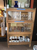Front view of oak Weis barrister bookcase showing three glass-front compartments filled with glasses and mugs (contents not included). Top covered with contact paper.