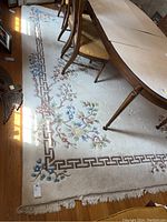 Rug under dining table showing floral motifs and Greek key border in cream and brown with fringed edges.