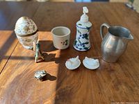 Full lot image showing eight items on wooden table including porcelain egg, milk glass hens, clay Pegasus, porcelain toad, pewter pitcher, soap dispenser, ceramic dish.