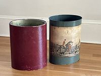 Photo showing two antique trash cans side by side against a white baseboard wall. The left can is burgundy leather-covered, round with metal interior. The right can is a metal cylindrical can with a faded painted hunting scene depicting horses and figures.