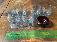 Seven liqueur glasses arranged with a measuring ruler for scale: Six greyish-clear rounded liqueur glasses with short stems and one small rounded ruby red glass cup.