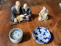 Photo showing two Hakata Urasaki dolls of elderly man and woman seated, a Napcoware figurine of a woman painting a pot, one OMC Japan plate, and a stack of six Arita Imari shallow bowls on a wooden table.