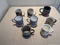 Eight different ceramic coffee mugs laid out on a beige cloth background showing various art styles and colors.