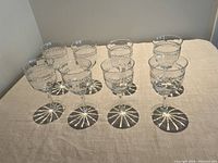 Eight clear crystal wine glasses arranged in two rows on a beige cloth. Each glass has an etched leaf pattern near the top rim and a round base. Light creates a distinct pattern of shadows beneath the bases.