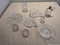 Full lot of nine assorted cut glass and crystal items on beige cloth including bowls, plates, jugs and bell.
