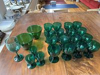 Overview of 20 emerald green glassware pieces arranged on wooden table. Various sizes and styles including wine glasses and bowls visible.