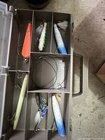Photo showing compartmentalized tackle box with 6 vintage lures including orange, white, green, and blue colors with feathered treble hooks and metal wire leaders.