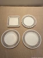Four milk glass plates arranged on a neutral fabric surface showing their lace-like pierced edge patterns, two round plates with twisted lattice edges, one round plate with looped lace edge, and one square plate with lattice border.