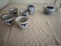 Four custard cups, one creamer and one sugar bowl arranged on a beige cloth; shows all items with floral and thistle designs.