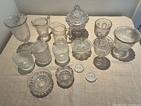 Top view of a broad collection of 18 vintage pressed glass pieces including pitchers, bowls, goblets, and small dishes displayed on light fabric surface.