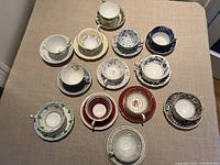 Overhead view of twelve bone china teacups with matching saucers arranged on beige cloth, displaying a variety of floral and decorative patterns across the collection.
