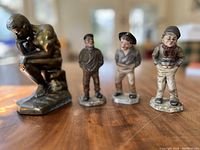 Photo showing the metal 'The Thinker' statue and three Royal Worcester porcelain street urchin figurines lined up on a wooden surface under natural light.