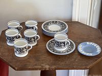 Photo showing the full set of six Wedgwood Florentine black and white demitasse cups and saucers arranged on a wooden table along with the single blue Wedgwood ashtray.