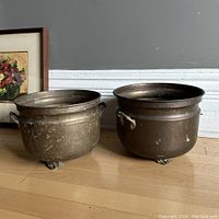 Front view of the two brass planter pots showing rounded shapes, handles, and claw feet with original patina and wear.