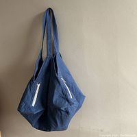 Blue square cotton tote bag hanging, showing two sides with visible zippered pockets.