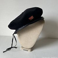 Side view of the black wool tam hat on beige mannequin showing leather brim trim and red enamel button.